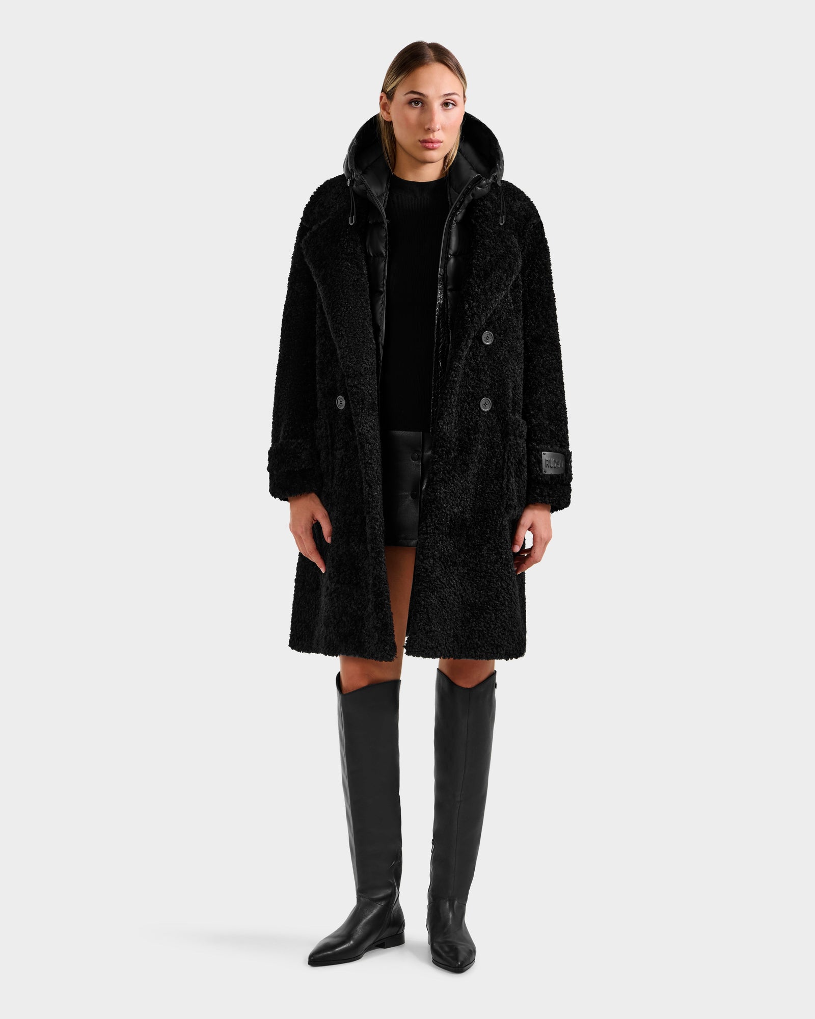A woman with light brown hair pulled back faces forward against a plain white background, wearing the Rudsak EMRYN - L125588 Black long winter coat over a black top and shorts, paired with knee-high black boots and a neutral expression.