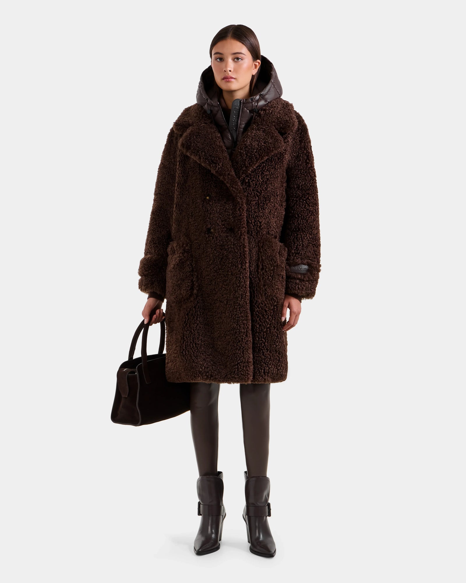 A woman with straight brown hair and a neutral expression wears the Rudsak EMRYN - L125588 Espresso long brown faux-fur winter coat over a black puffer jacket, brown leggings, black ankle boots, and holds a black handbag.