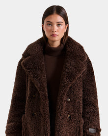 A woman with straight brown hair wears Rudsak’s EMRYN - L125588 Espresso teddy jacket over a brown turtleneck, standing against a plain, light background.