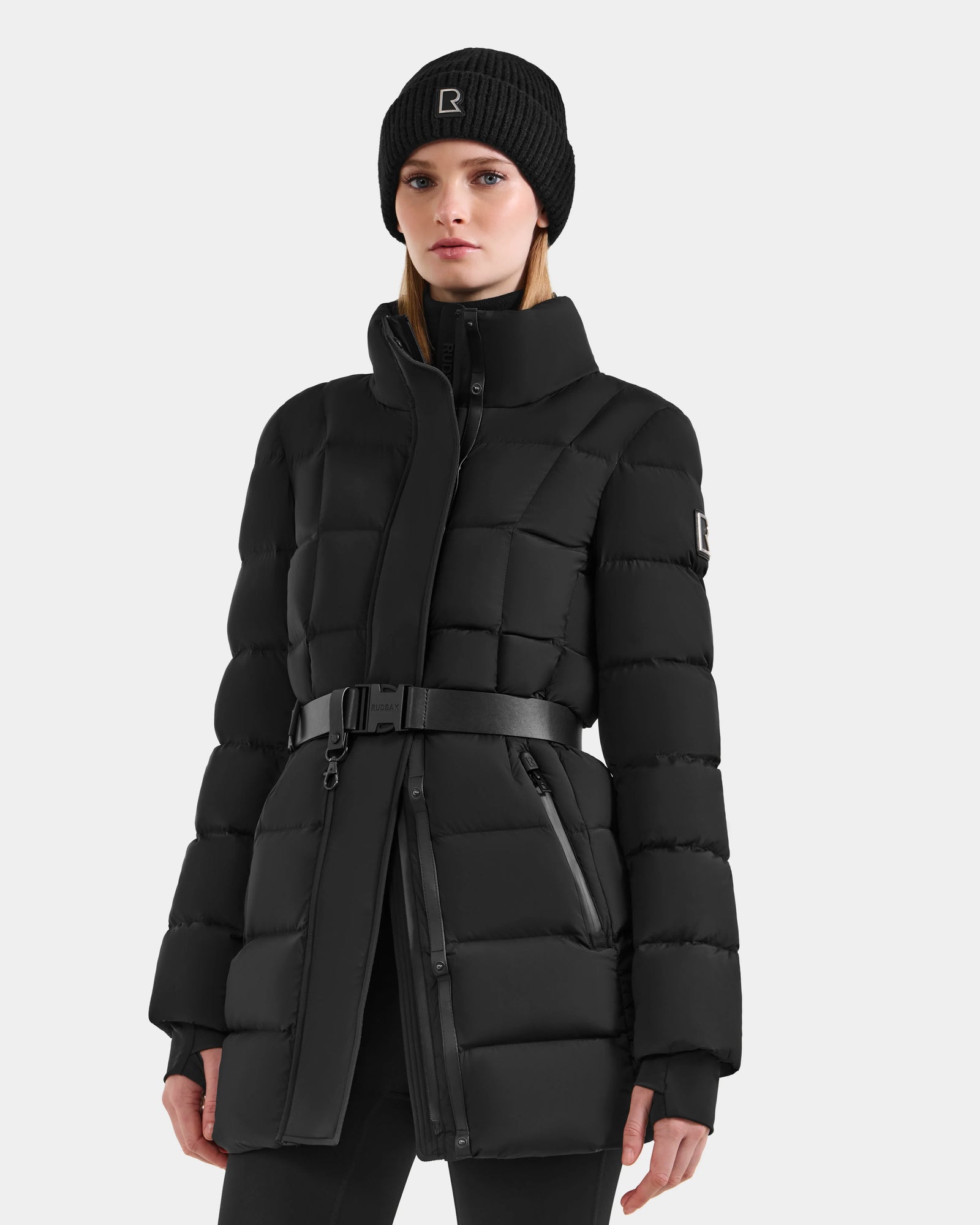 A woman wears the Rudsak FRIDA M-M - L125607 Black quilted down puffer coat with belted waist and detachable hood, plus a matching black knit beanie, standing against a plain light background—your versatile winter essential.