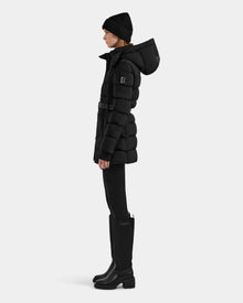 A person in profile wears the Rudsak FRIDA M-M - L125607 Black quilted down puffer with a detachable hood, styled with a black knit beanie, leggings, and tall boots against a light gray background—a chic winter essential.