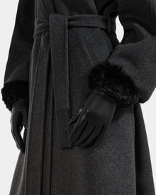 A close-up of a person wearing the Rudsak GERRY - L425550 Black belted coat and black faux-fur cuff gloves with a Rudsak tag. The women’s leather gloves add elegance against the plain white background.