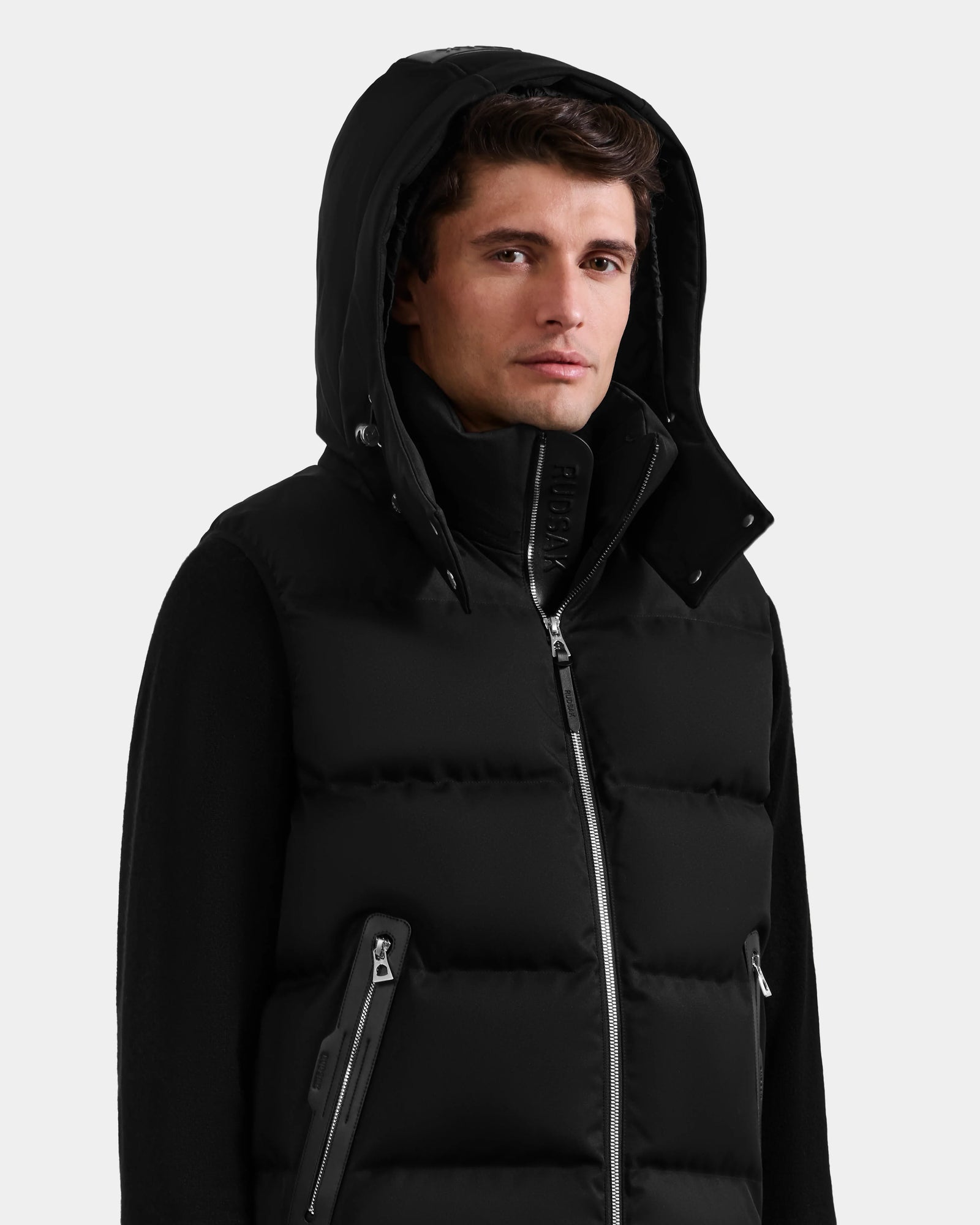 A young man wearing the Rudsak HARLOW - M125528 Black men’s puffer vest with a high collar, zippered pockets, and a removable hood stands against a plain light gray background.