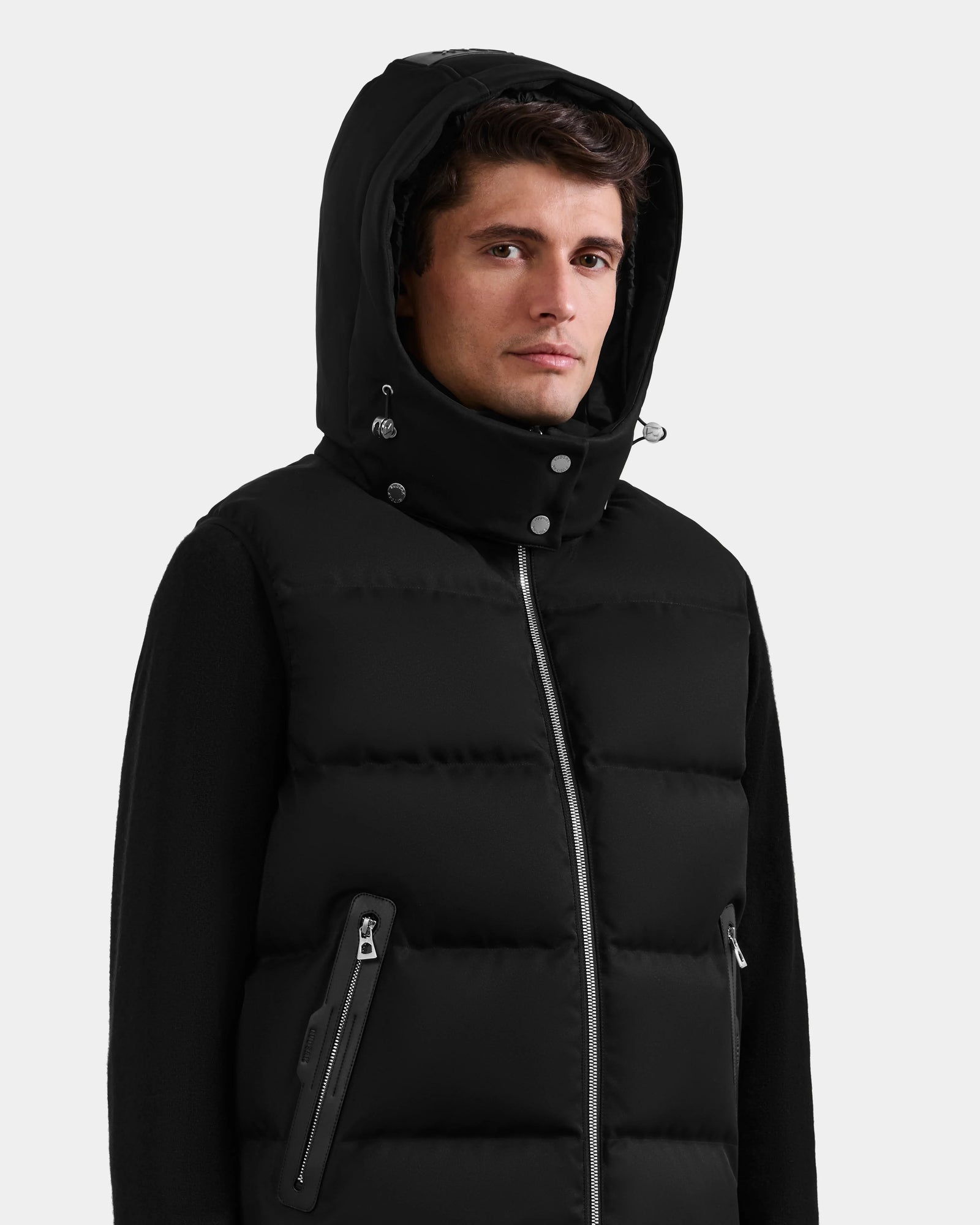 A man wears the Rudsak HARLOW - M125528 Black quilted down vest with zipper, zippered pockets, and a removable hood, standing against a plain light gray background.