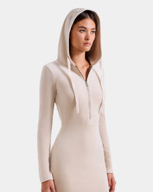 A woman wears the Rudsak ISABELLA - L525502 Cream dress, a fitted light beige velour hooded style with long sleeves, front zipper, and subtle RUDSAK logo, standing against a plain white background.
