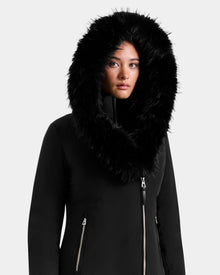 A woman wearing the Rudsak JANIS - L125558 Black parka with a large, fluffy black fur hood looks forward with a neutral expression against a plain light background—a stylish cold-weather layer.