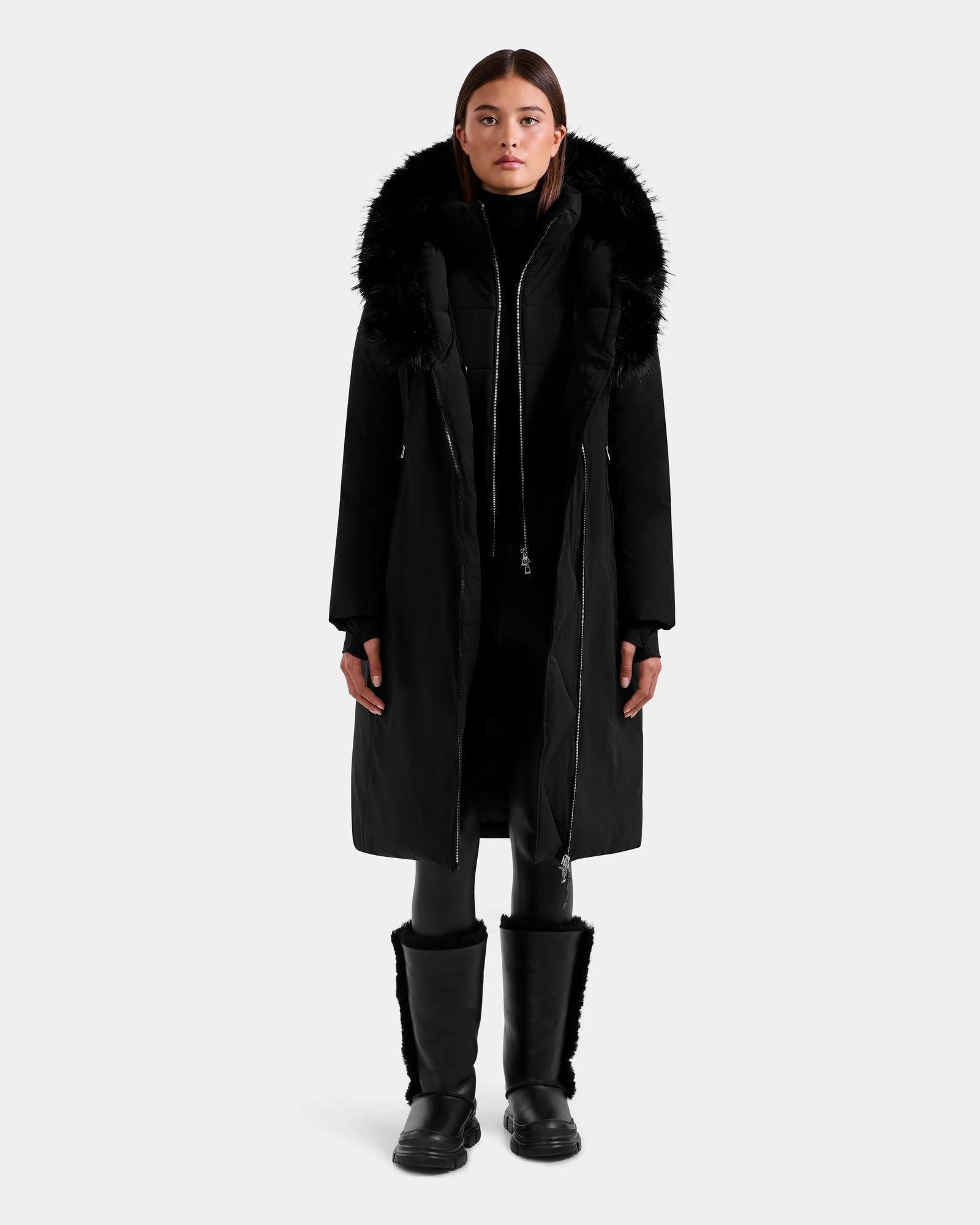 A person faces forward against a plain light background wearing the Rudsak JANIS - L125558 Black long winter coat with a faux fur hood, styled with a black turtleneck, leggings, and boots.