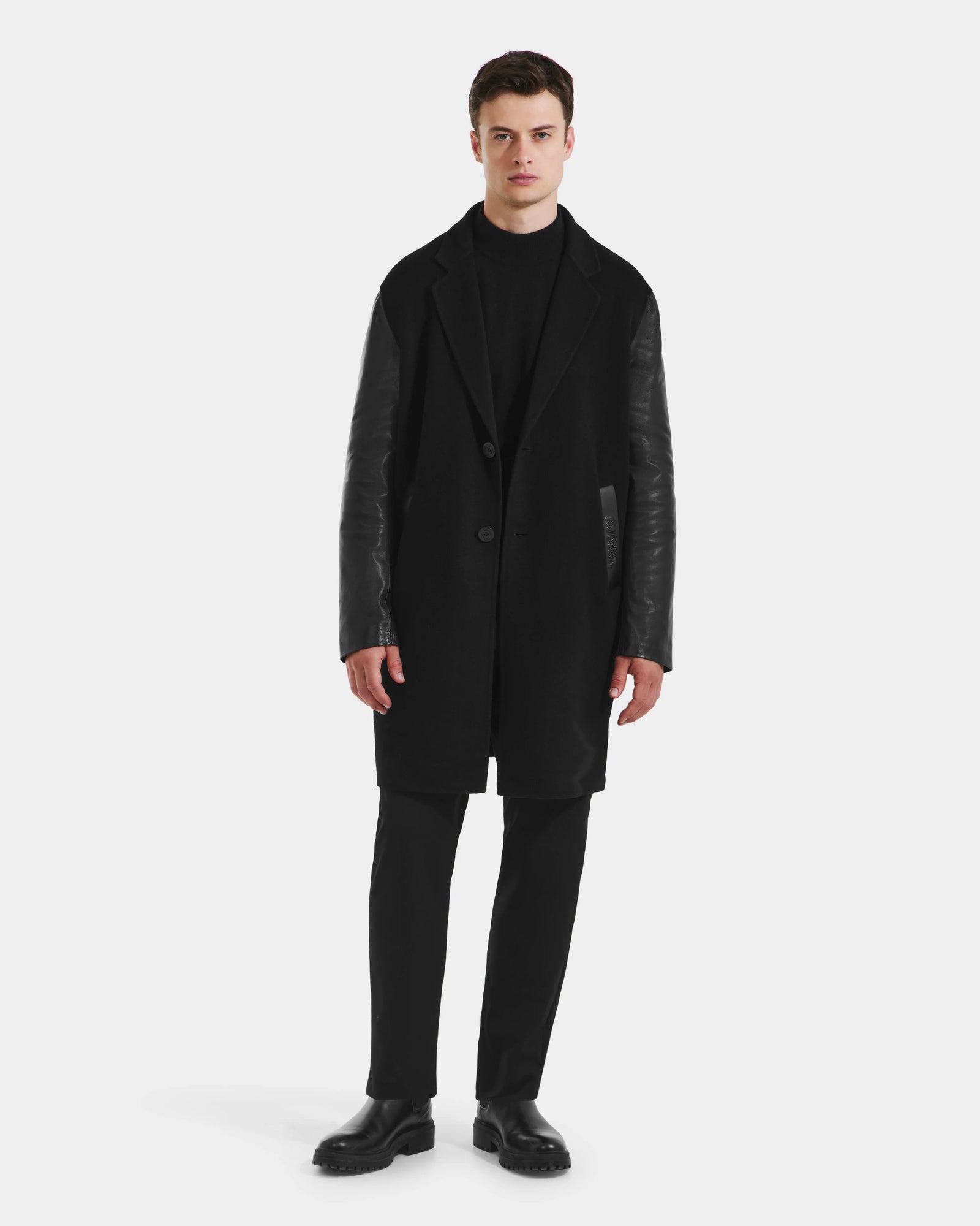 A man stands against a white background wearing the Rudsak JESSE - M125504 Black men’s wool coat with leather sleeves, paired with black pants and shoes. He faces forward neutrally, arms at his sides.