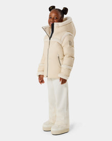A young girl with braided hair smiles in the Rudsak JULIANA K - K125550 Vanilla puffer jacket, featuring faux shearling trim, paired with white pants and cream platform shoes against a plain white background.