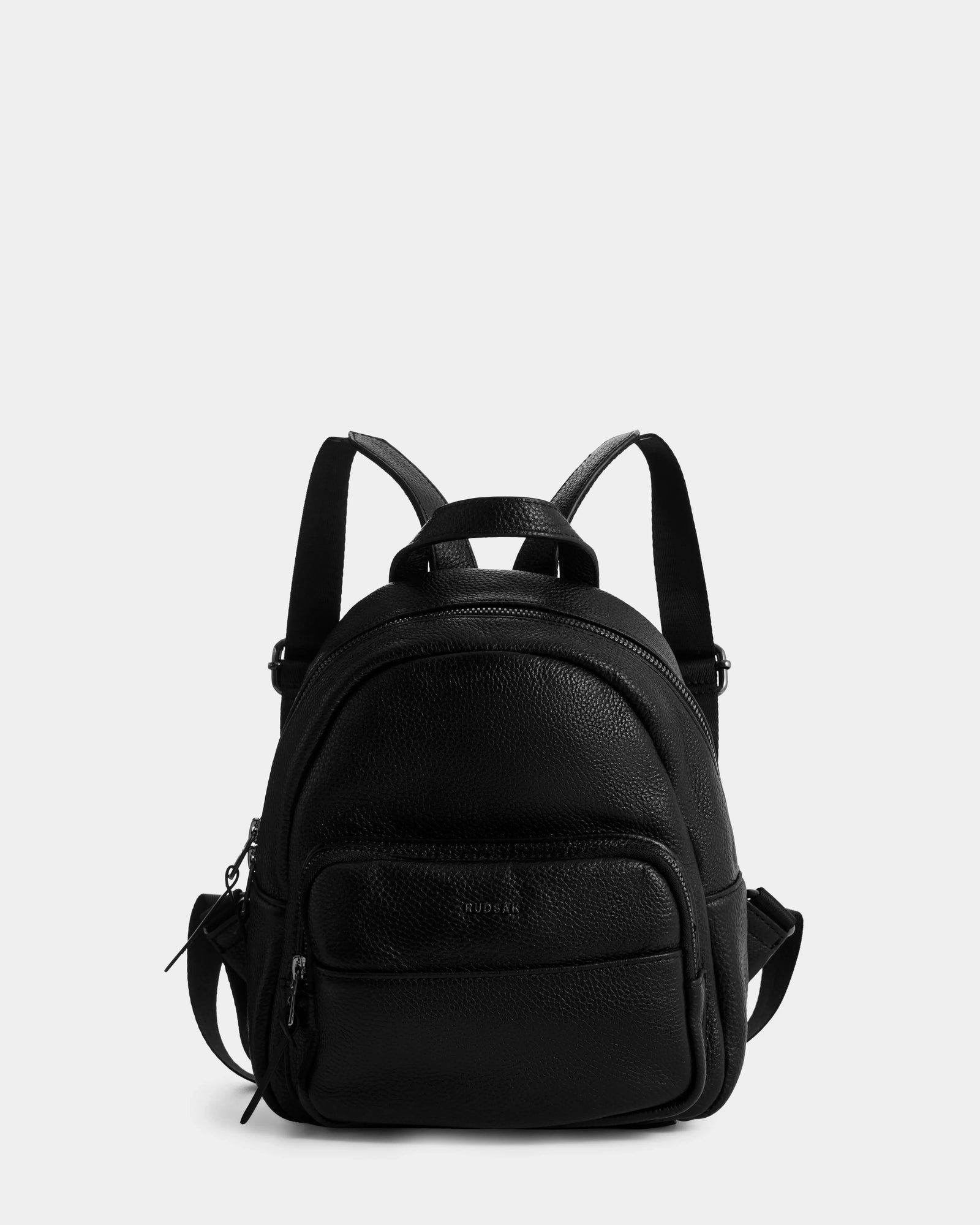 The Rudsak KATTY - L325014 Black is a women’s mini backpack in black pebbled leather with adjustable straps, a front zip pocket, and main compartment, shown on a plain light gray background.