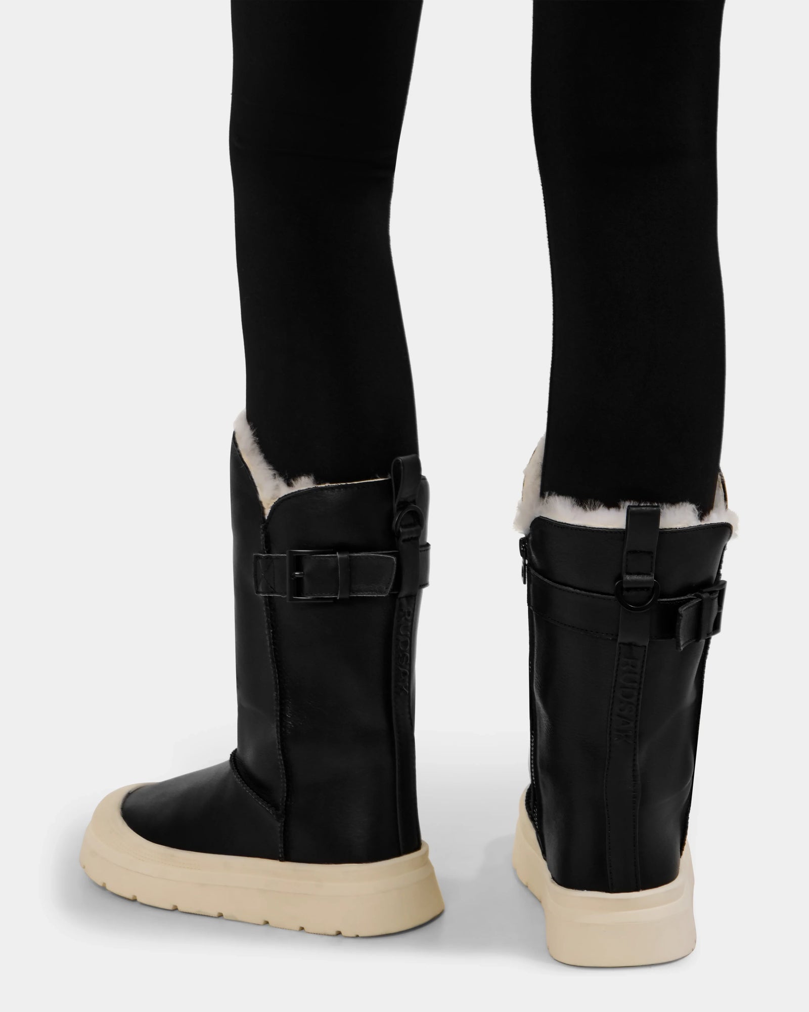 Model wears the Rudsak KLAIR L - L225519 Black boots, featuring thick light-colored soles, city-ready traction, white fleece lining, side buckle straps, and vertical textured back detail. Paired with black leggings.