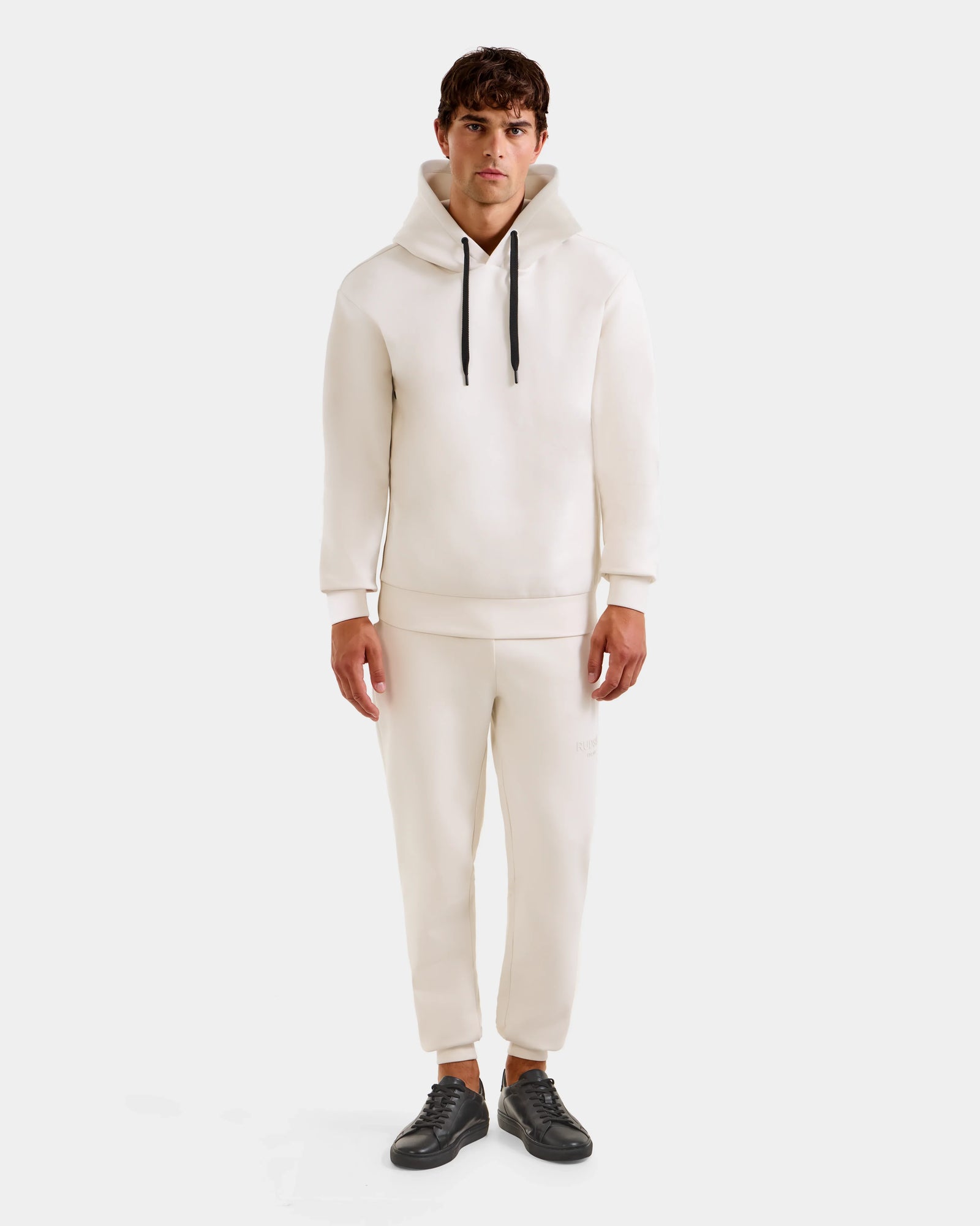 A man faces forward in a Rudsak LEROY - U525509 Vanilla M cotton-blend hoodie with black drawstrings, paired with matching off-white jogger pants and black sneakers, set against a plain white background.