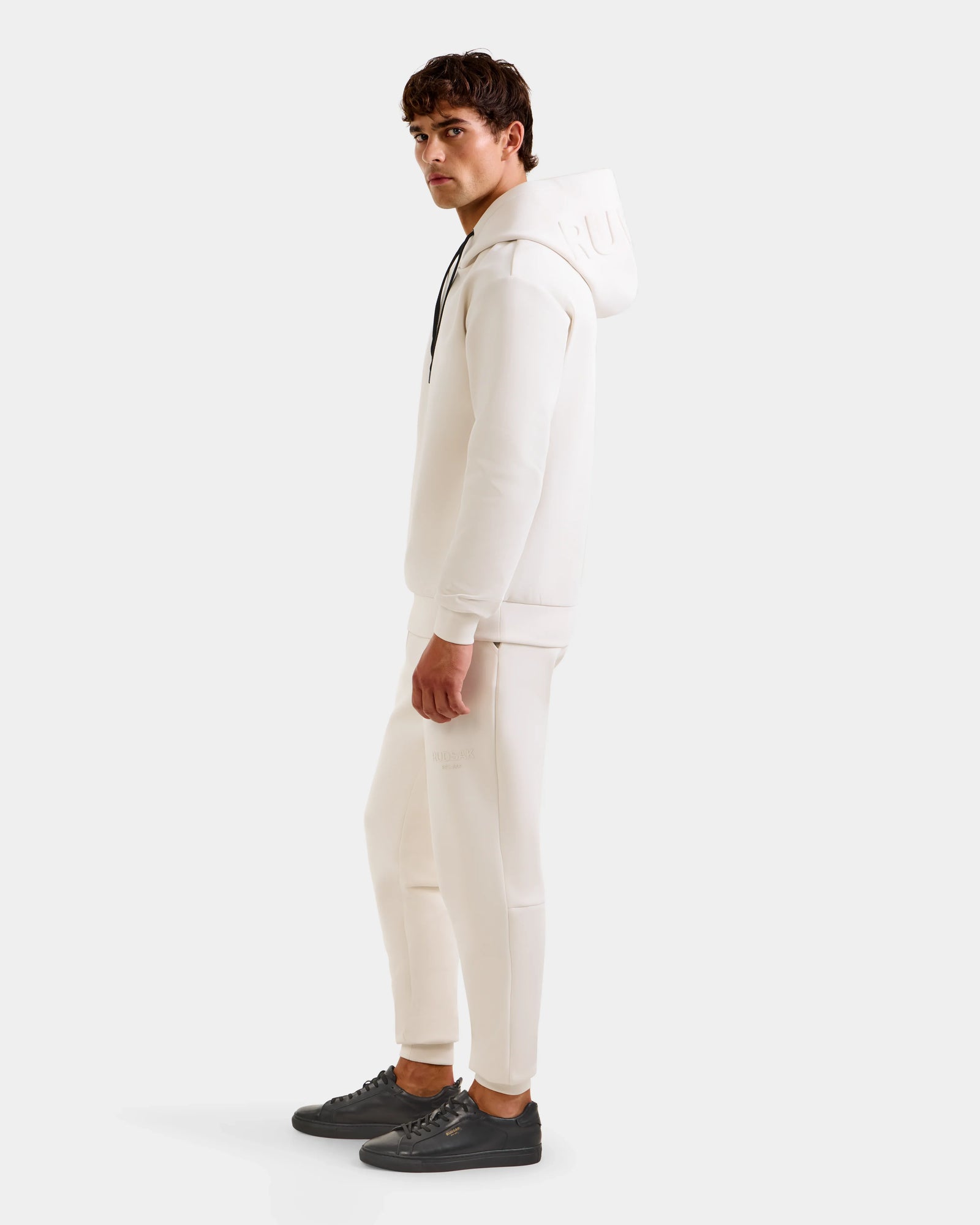 A man stands sideways against a plain light background, wearing the Rudsak LEROY - U525509 Vanilla M premium unisex hoodie, matching white pants, and black sneakers.