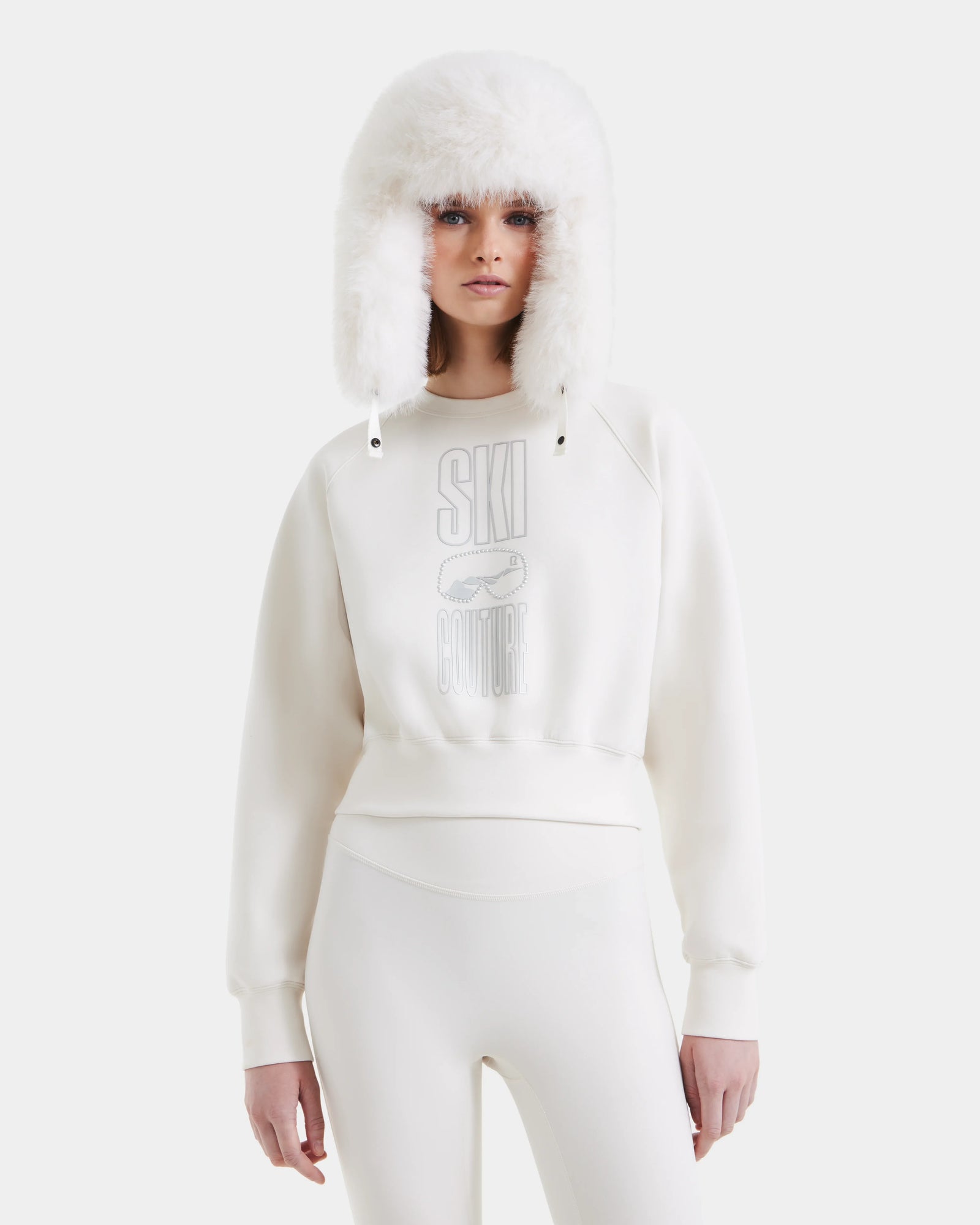 A woman stands against a white background wearing the Rudsak LIDDIE - L525505 Vanilla women’s cotton-blend sweatshirt with SKI COUTURE on the front, styled with fitted pants and a fluffy trapper hat for chic après-ski flair.