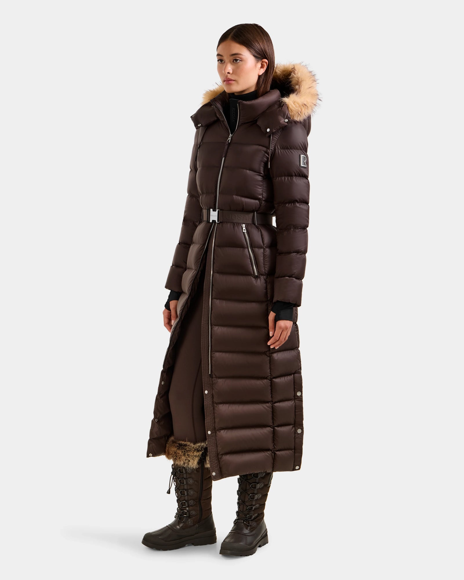 A woman stands against a plain background wearing the Rudsak LILITH L - L125528 Espresso maxi-length down puffer coat with a fur-trimmed hood, removable belt, black gloves, dark pants, and brown lace-up winter boots with fur lining.