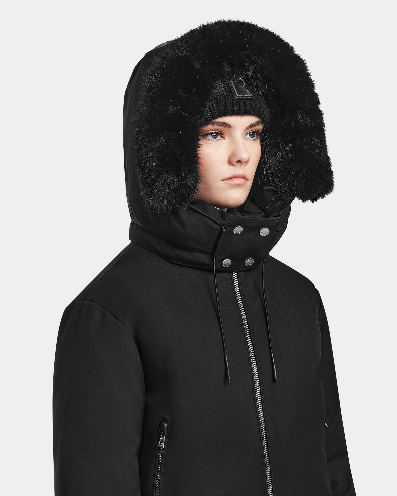 A person wears the Rudsak MAUDE - L124522 Black/Black women’s down bomber with a large fur-lined hood and a black beanie, looking ahead with a neutral expression against a plain background.