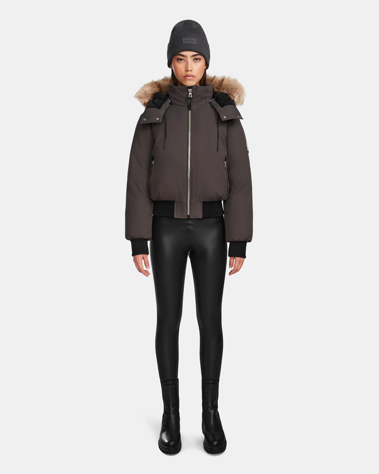 A woman faces forward against a white background, wearing the Rudsak MAUDE - L124522 Grey jacket—a dark brown puffer with premium down and fur-lined hood—paired with a gray beanie, black leggings, and black boots.