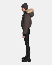 A person stands in profile against a plain white background, wearing the Rudsak MAUDE - L124522 Grey women’s down bomber with fur-lined hood, paired with a gray beanie, black leggings, and black boots.
