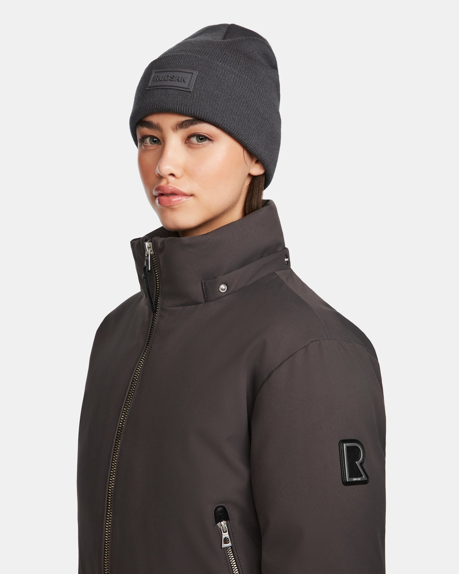 A young woman models the Rudsak MAUDE - L124522 Grey beanie and a dark high-collar winter jacket, standing against a plain white background and gazing slightly to the side.