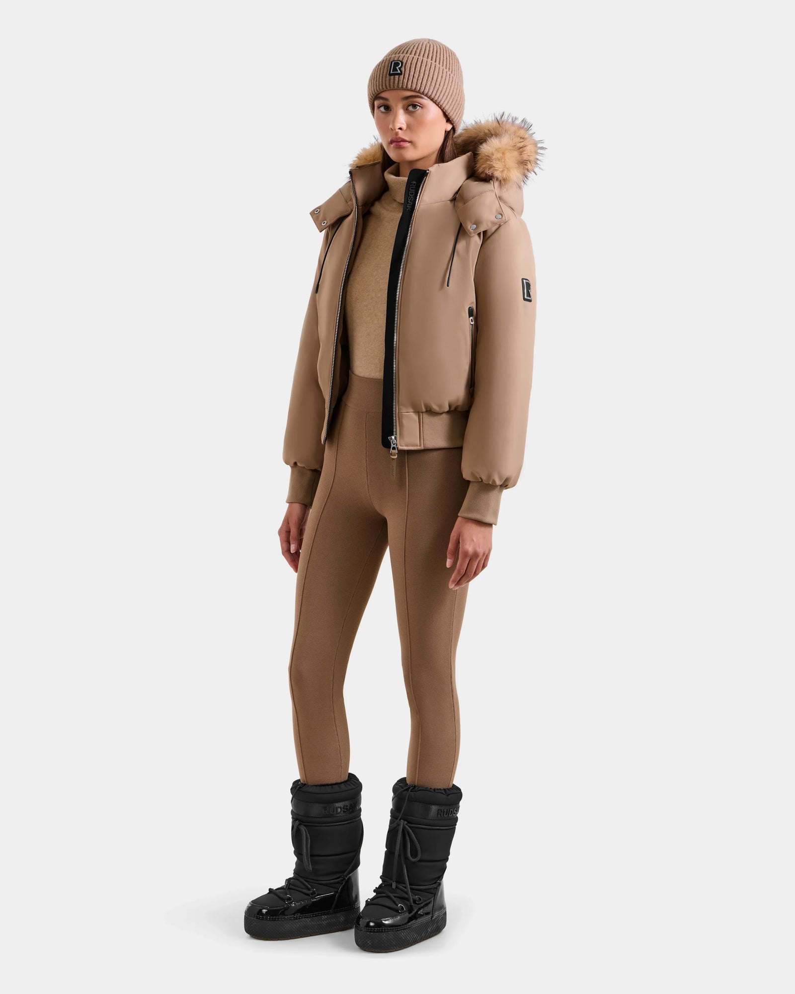 A woman stands against a plain white background wearing the Rudsak MAUDE - L124522 Sand women's down bomber with a fur-trimmed hood, tan knit beanie, fitted pants, and black winter boots.