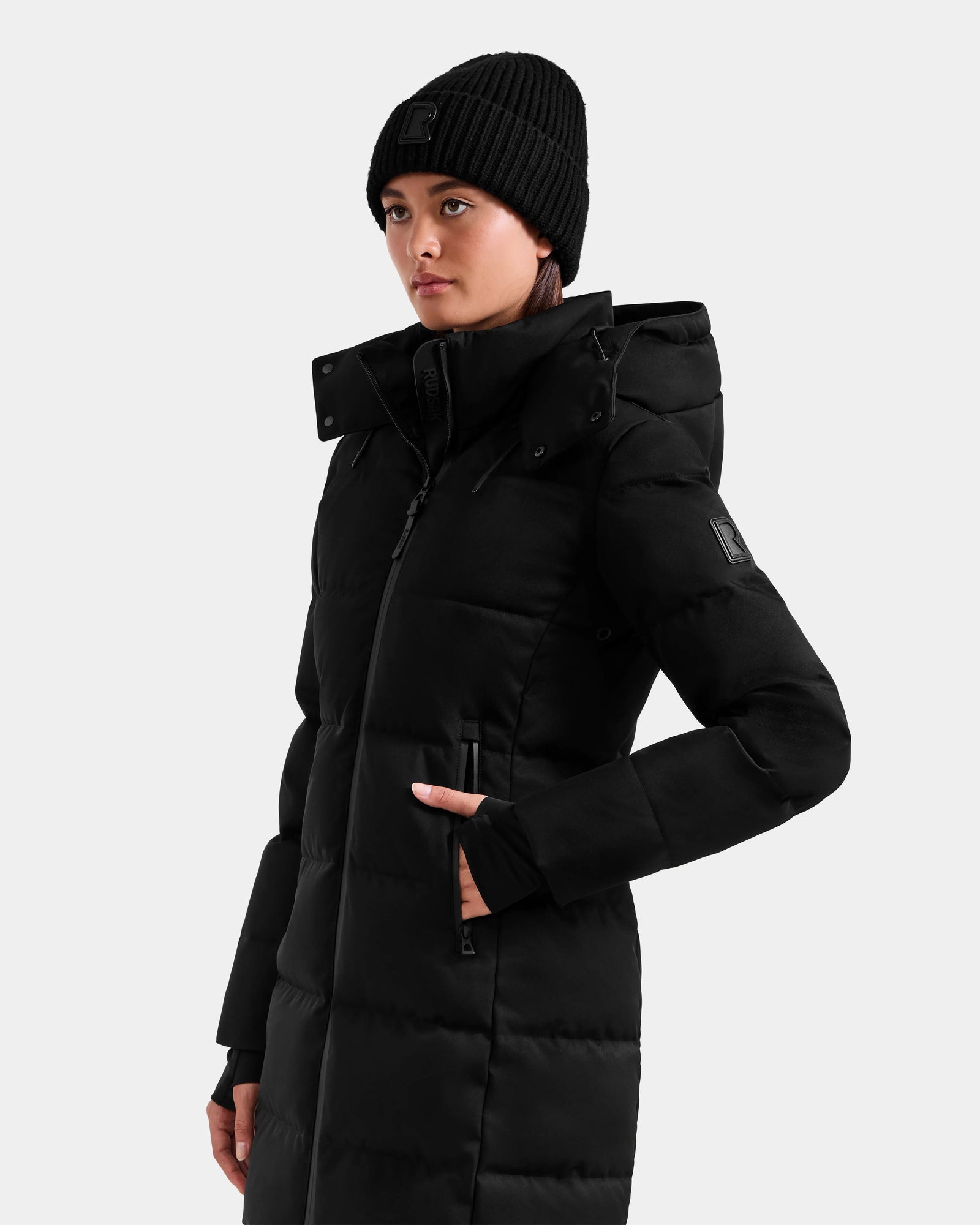 Wearing the Rudsak MIRIAM MW - L125604 Black women’s down puffer coat with a hood and a black knit beanie, she stands against a plain light background, looking slightly to the side with one hand in her pocket.