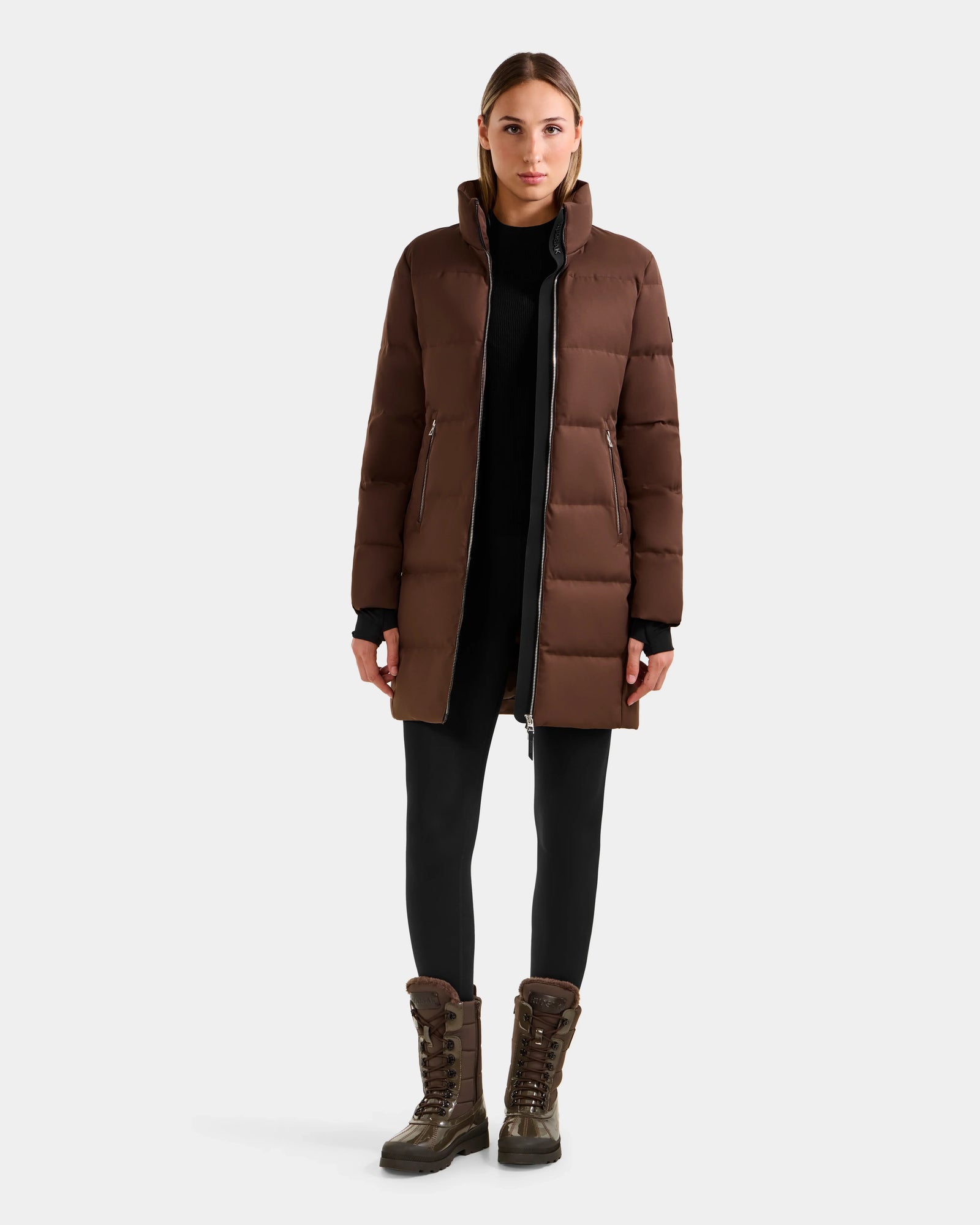 A woman faces forward against a light background, wearing the Rudsak MIRIAM - L123544 Mocha long down puffer coat, paired with black leggings and brown lace-up boots. Her straight hair is pulled back and her hands rest by her sides.