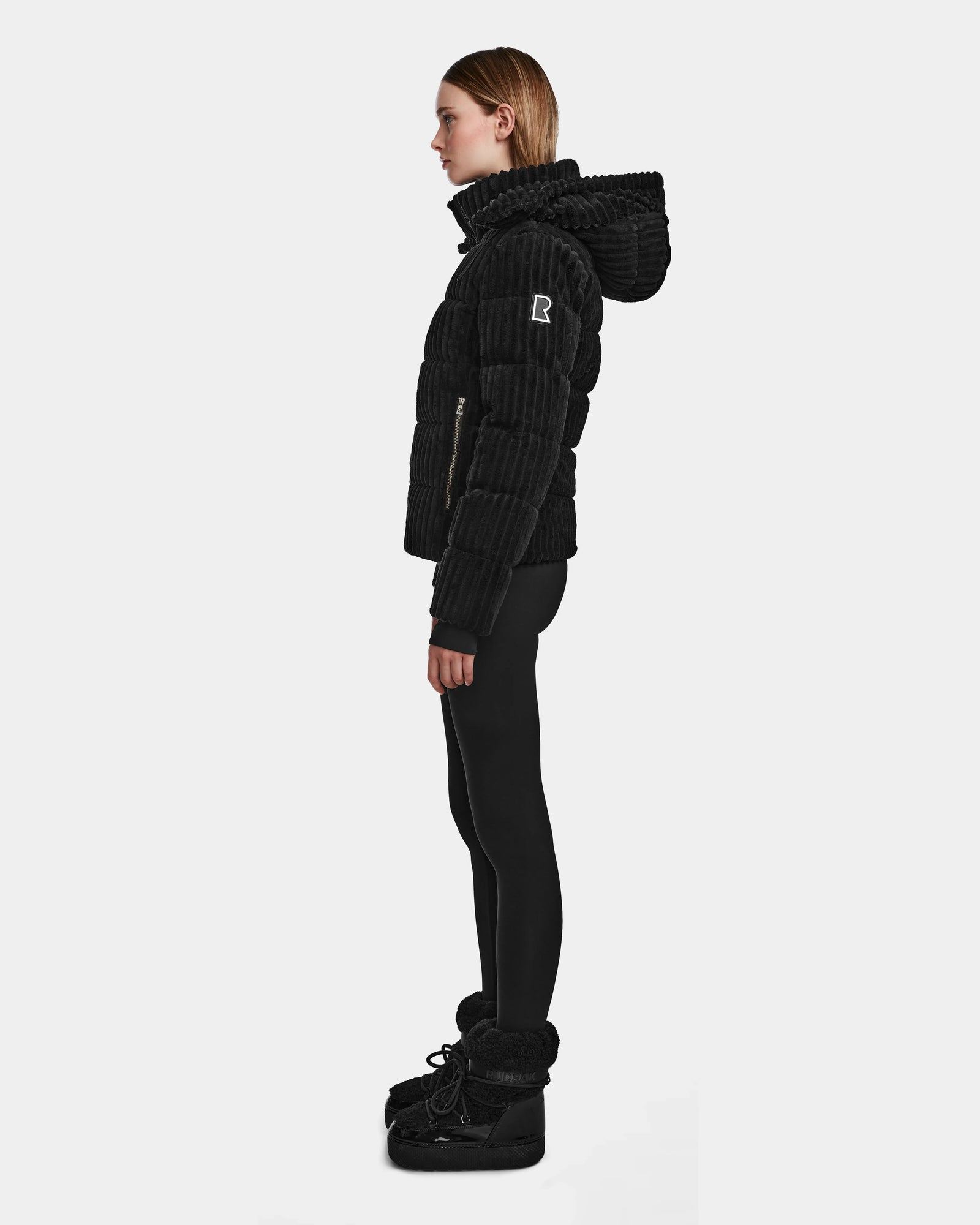 A woman stands in profile wearing the MIRRY FF - L124507 Black women’s short down puffer jacket with a hood, black leggings, and black winter boots against a plain white background. The jacket features RDS-certified down for warmth and ethics.