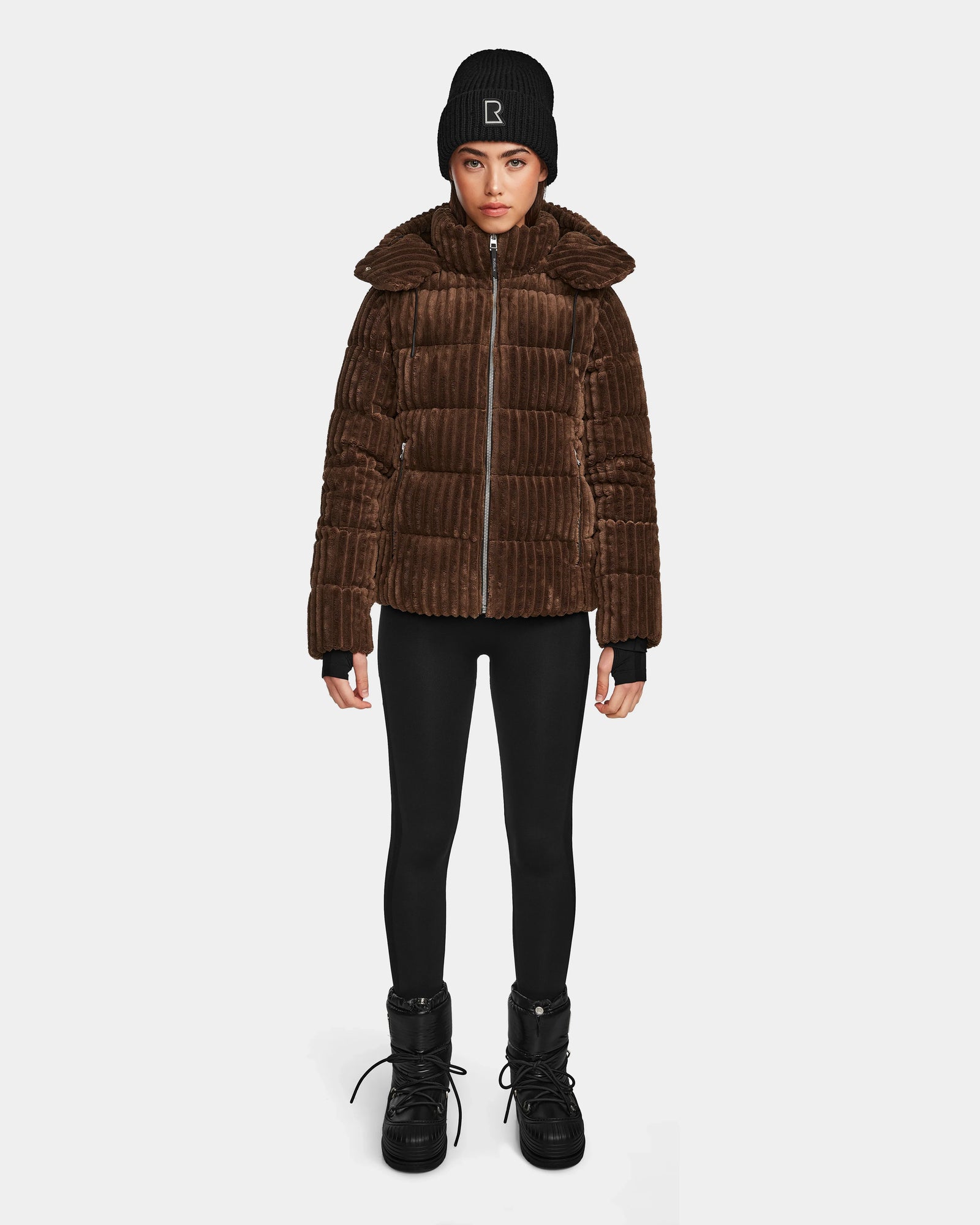 A person stands facing forward wearing the Rudsak MIRRY FF - L124507 Espresso women's short down puffer jacket, paired with black leggings, black lace-up boots, and a black beanie against a plain white background.