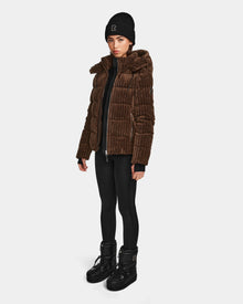 A woman stands against a white background wearing the Rudsak MIRRY FF - L124507 puffer jacket in Espresso, paired with black leggings, winter boots, and a black beanie. She looks forward with a neutral expression.