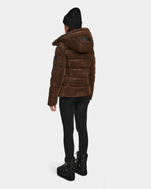 A person wears the Rudsak MIRRY FF - L124507 Espresso velvet corduroy puffer jacket, paired with a black beanie, leggings, and boots, standing with their back to a plain white background.