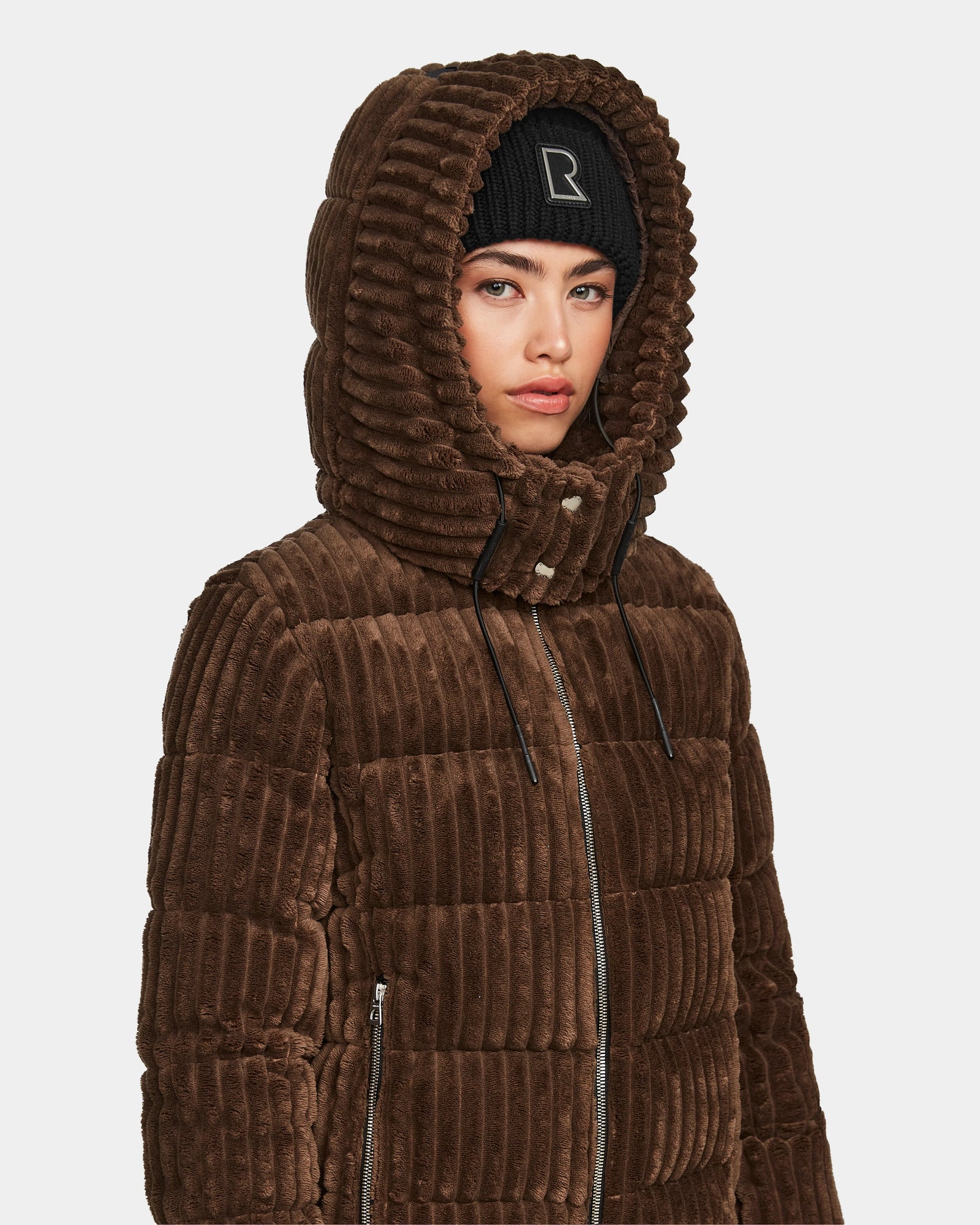 A woman wears the Rudsak MIRRY FF - L124507 Espresso short down puffer jacket with a hood and black beanie. The brown, corduroy-style quilted coat features a zippered front. She stands slightly turned against a plain background.
