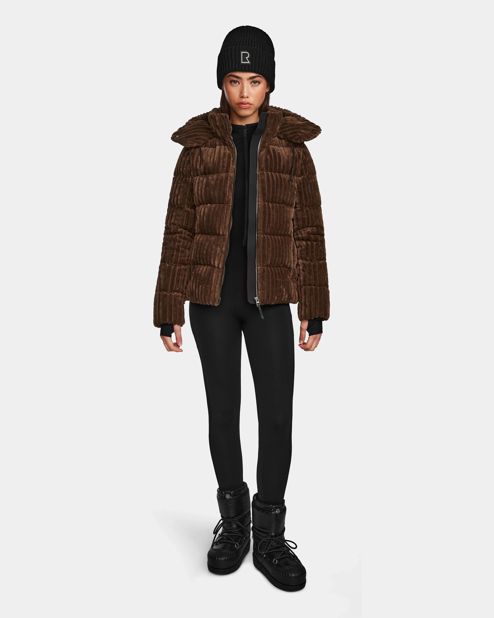 A woman faces forward against a white background, wearing the Rudsak MIRRY FF - L124507 Espresso women’s short down puffer jacket, black leggings, black winter boots, and a black beanie with a white "R".