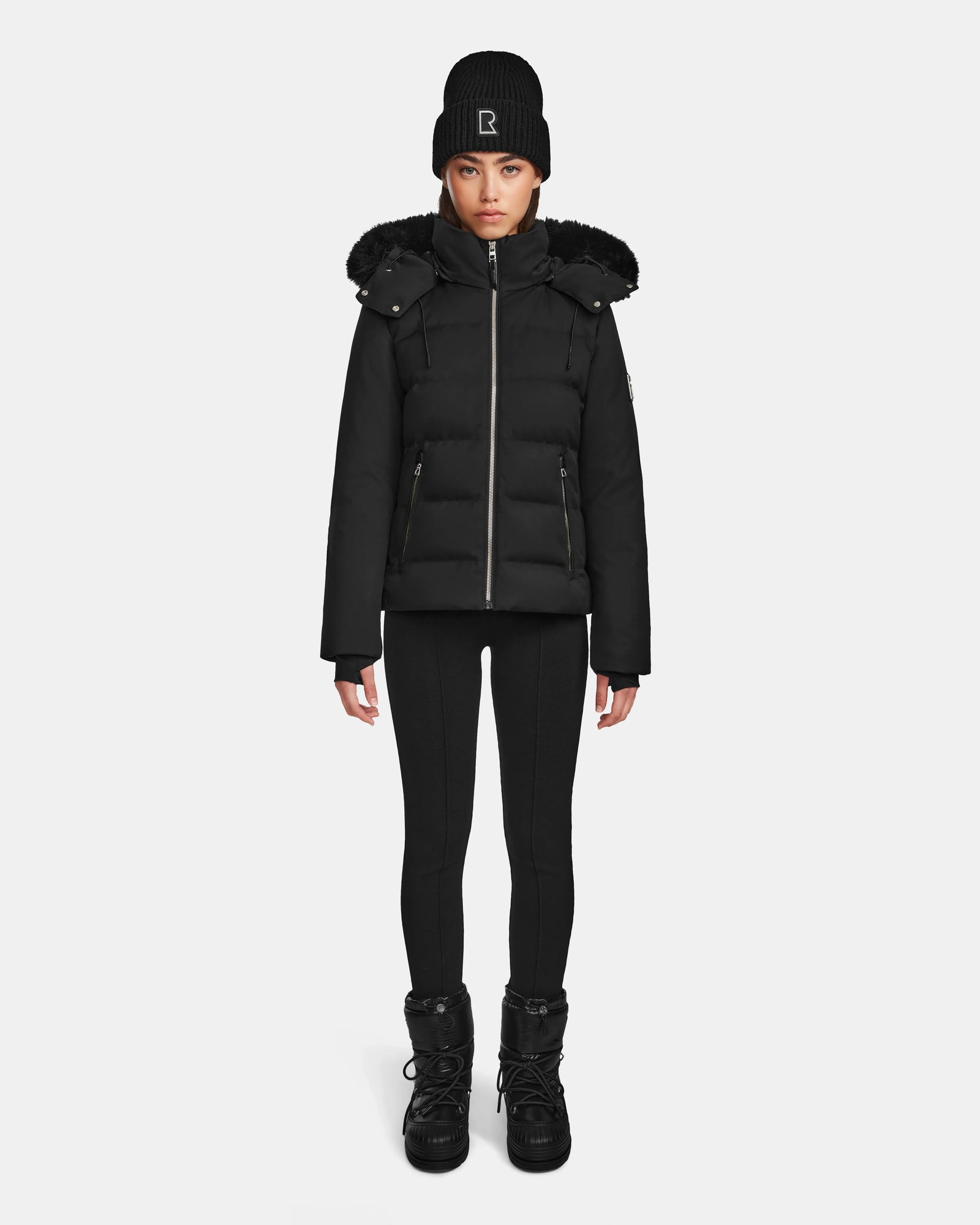 A person stands facing forward against a plain white background, wearing the Rudsak MIRRY M - L124533 Black women’s quilted down coat with fur-trimmed hood.