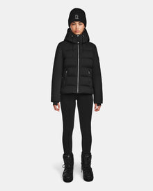 A person stands facing forward against a plain white background, wearing the Rudsak MIRRY M - L124533 Black puffer jacket, black leggings, black winter boots, and a black knit beanie with a small white logo.