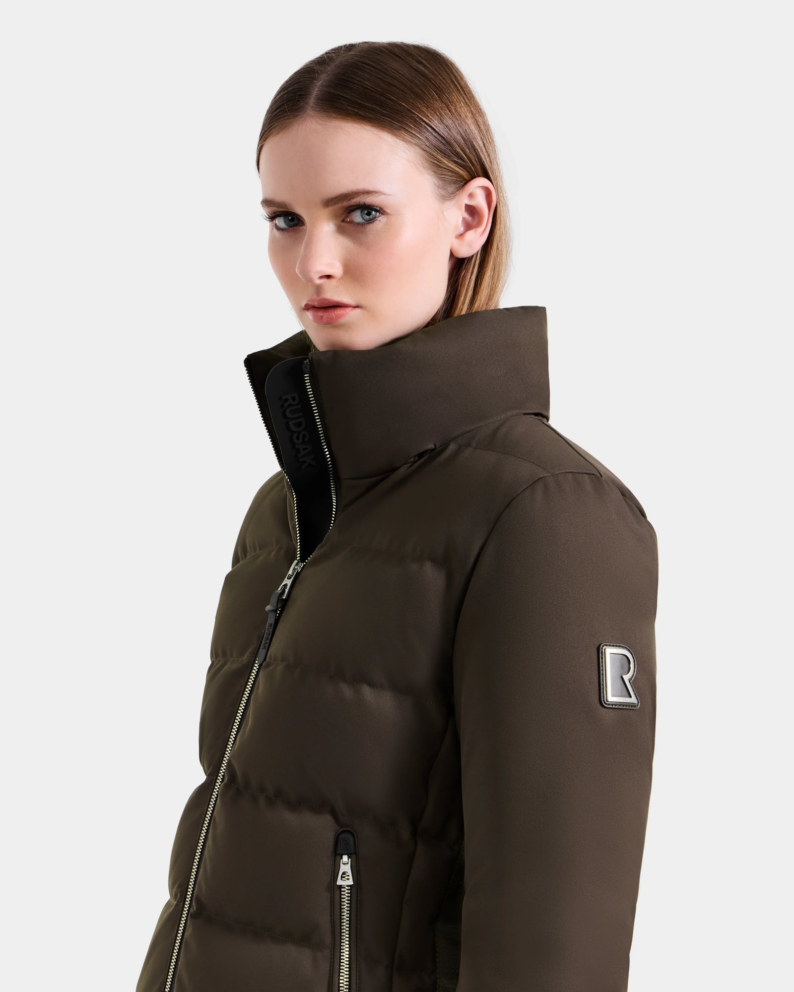 A woman with straight, light brown hair wears the Rudsak MIRRY M - L124533 Espresso high-collared, dark olive green quilted down coat with a zipper and an "R" patch on the sleeve, standing against a plain light background.