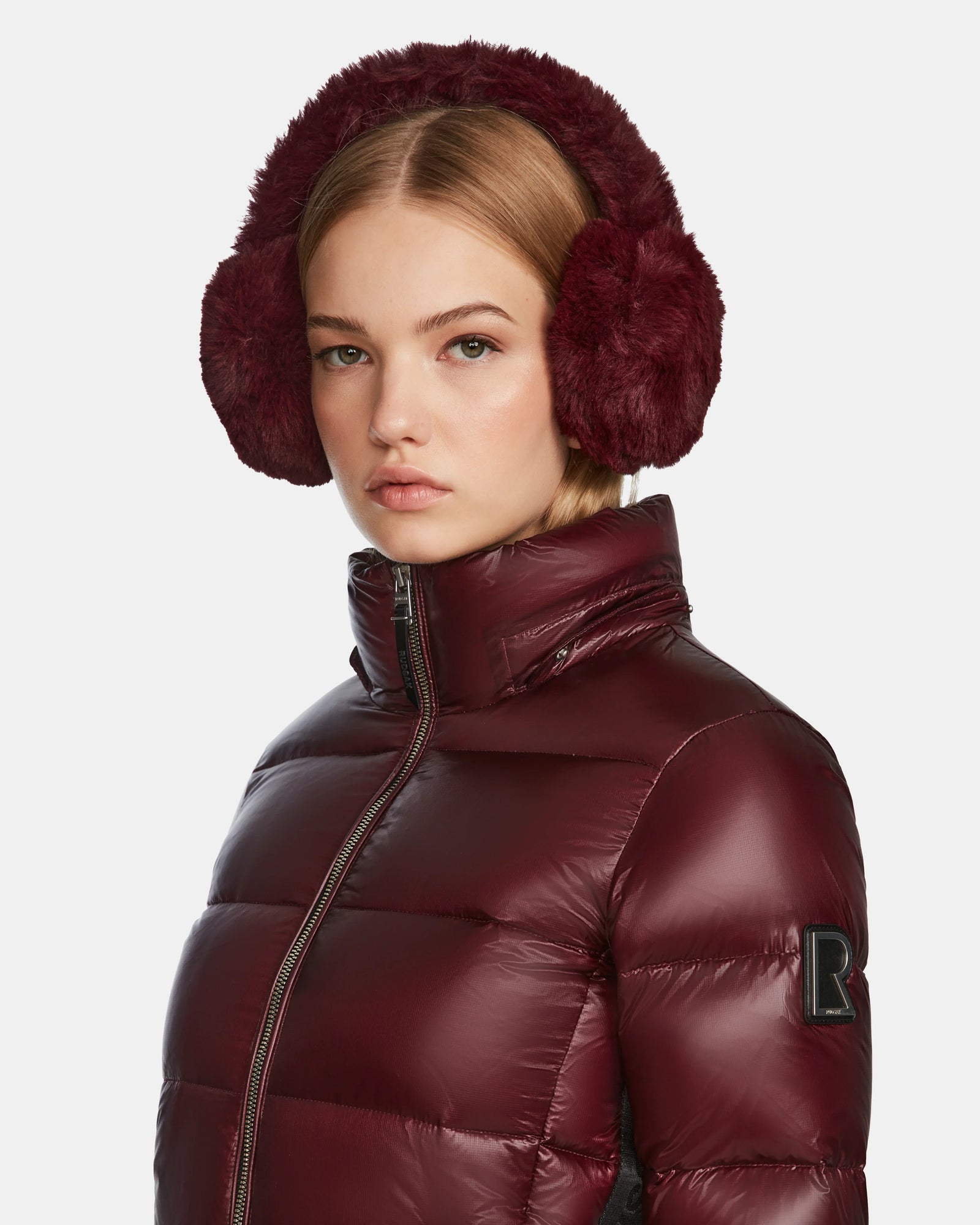 A young woman wears burgundy faux fur earmuffs and the Rudsak MIRRY S - L123545 Wine womens quilted down coat with a high collar, gazing forward with a neutral expression against a plain background.