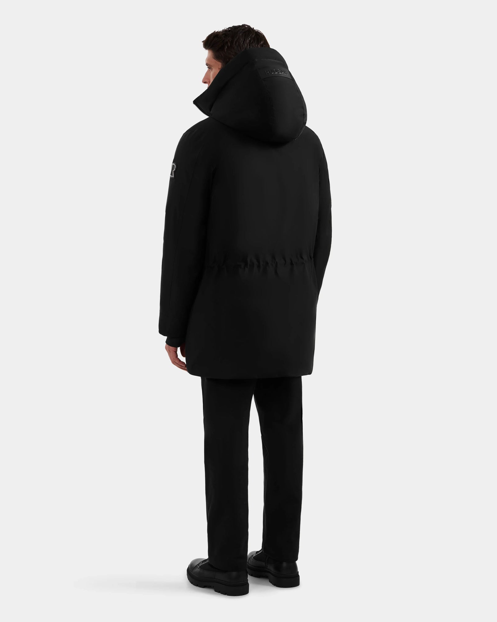 A man faces away wearing the Rudsak MITCHELL - M125513 Black men's down parka, paired with black pants and shoes, set against a plain light gray background.