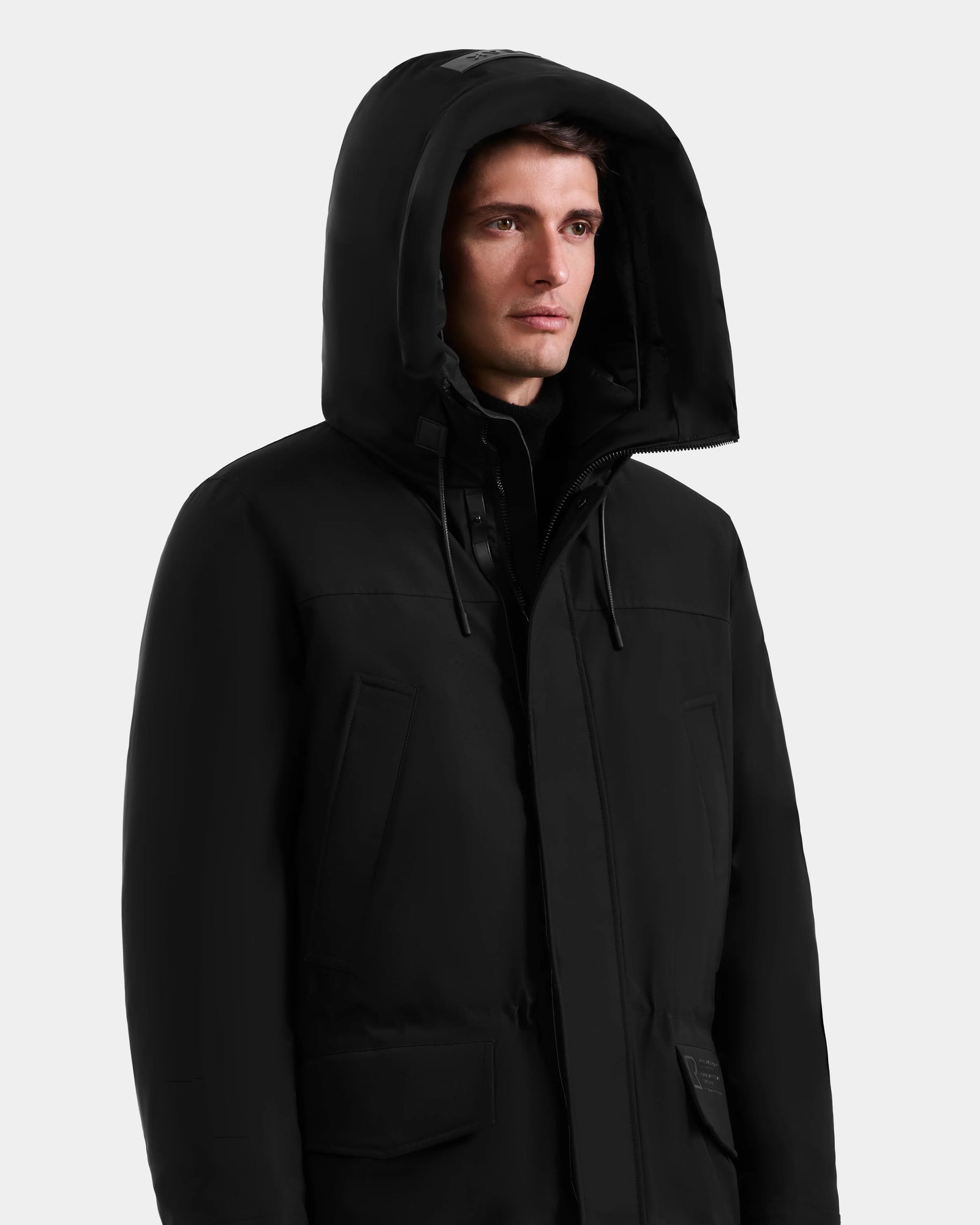 A man wearing a black Rudsak MITCHELL - M125513 down parka stands against a plain light gray background, looking slightly to the side with a neutral expression.