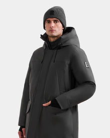 A man wears the Rudsak MITCHELL - M125513 Grey men’s down parka, featuring RDS-certified down, a sleeve patch, and zippered front pockets. He pairs it with a gray knit beanie, looking to the side against a plain light background.
