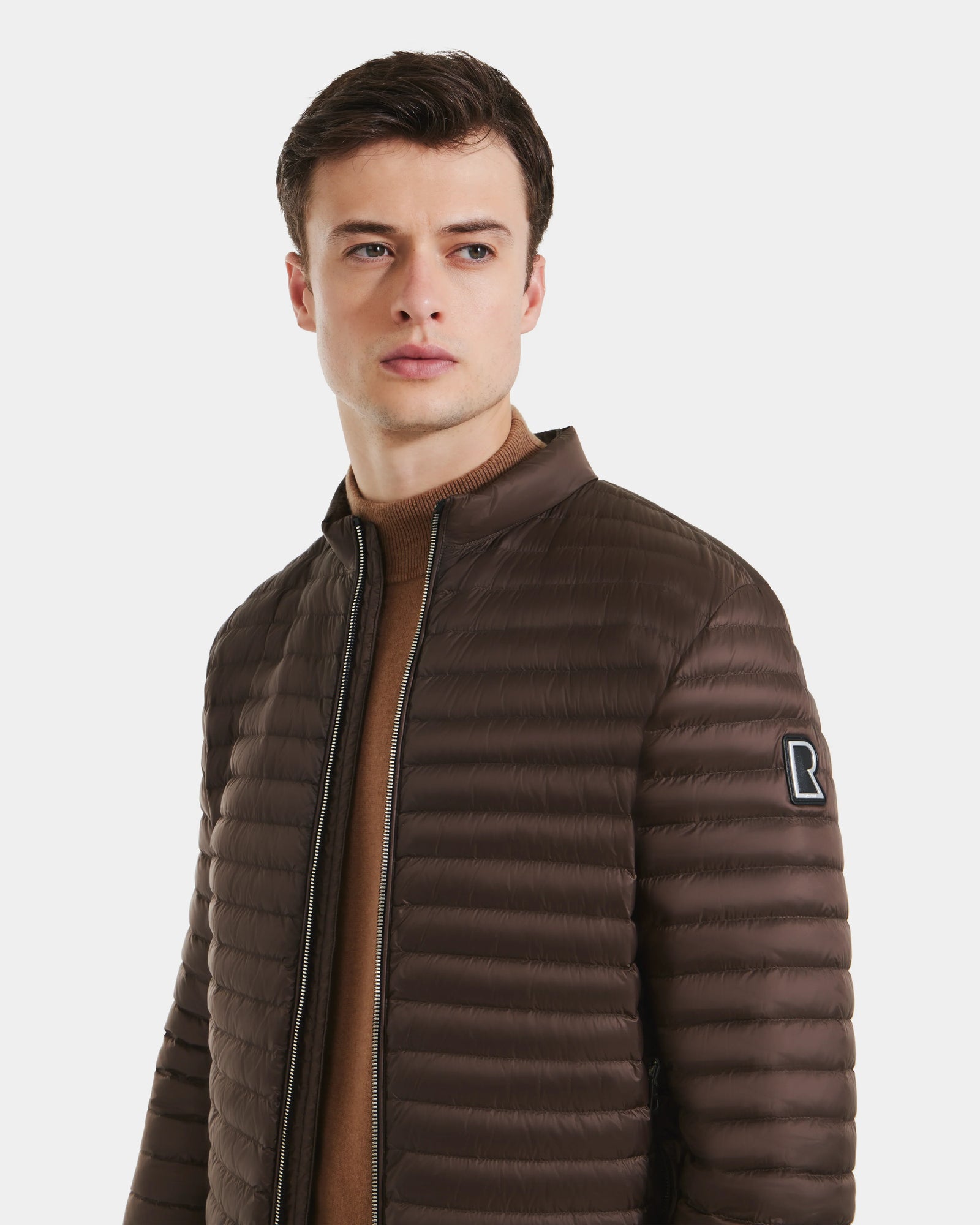 A young man with short dark hair wears the Rudsak REYMOND M - M125523 Mocha quilted men’s down jacket filled with RDS-certified down over a tan sweater, looking slightly to the side against a plain white background.