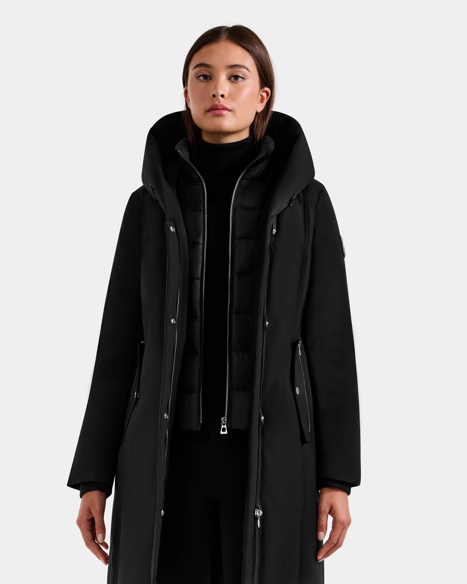 A woman models the Rudsak SARINA - L125550 Black parka, featuring a long semi-fitted design, high collar, and silver zipper. She stands against a plain light background with straight shoulder-length brown hair and a neutral expression.