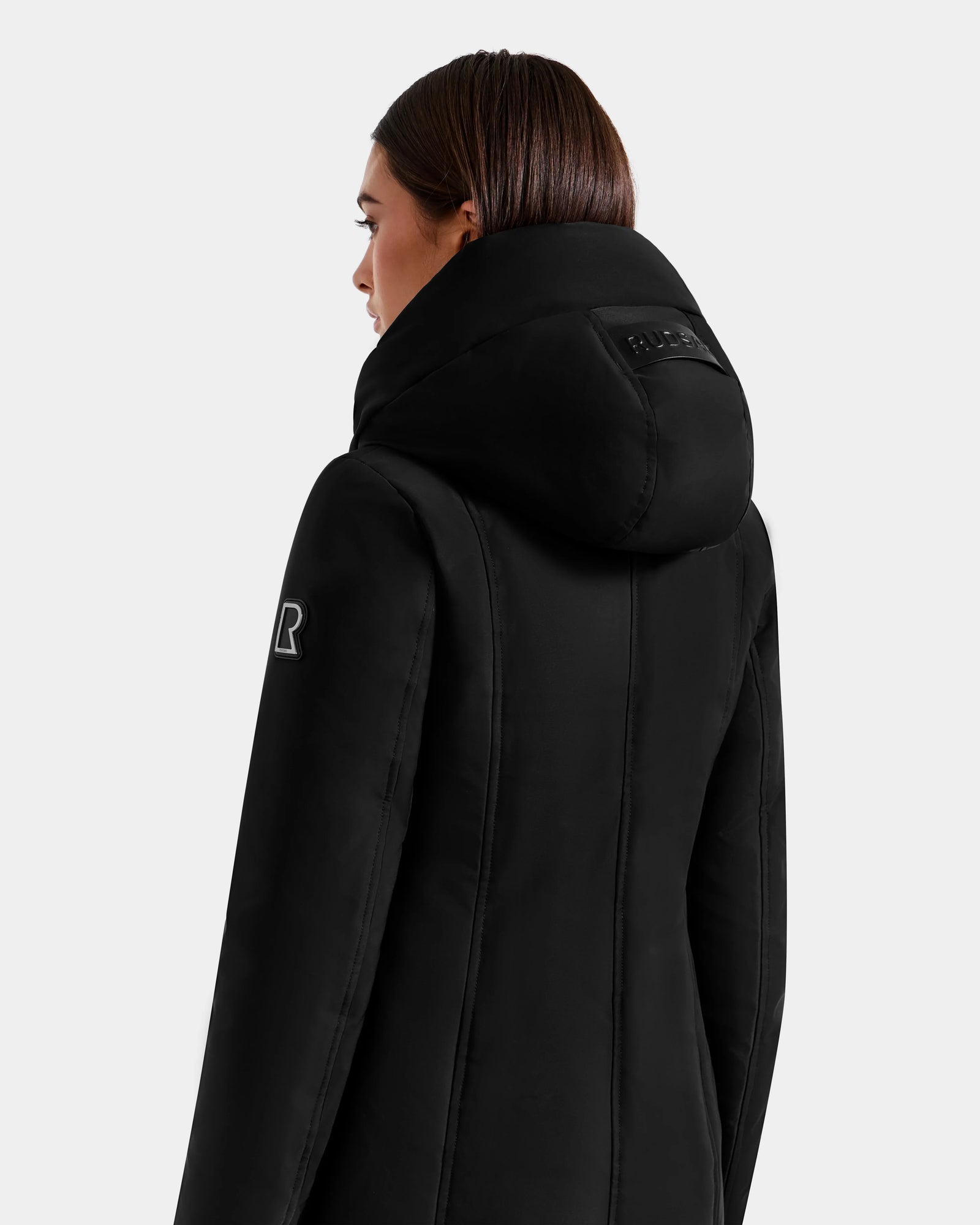 A woman with straight brown hair models the Rudsak SARINA - L125550 Black women’s down parka, shown from behind and in profile against a plain light gray background.