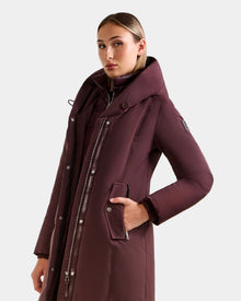 A woman models the Rudsak SARINA - L125550 Bordeaux: a long women's down parka with a high collar and zipper details, filled with RDS-certified down. She stands confidently, one hand in her pocket, against a plain light background.