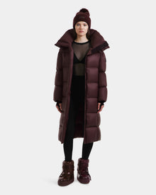 A woman stands against a white background wearing the Rudsak SASHA - 8125686 Bordeaux women’s long down puffer coat, a matching maroon knit beanie with pom-pom, sheer black top, black leggings, and maroon high-top boots.