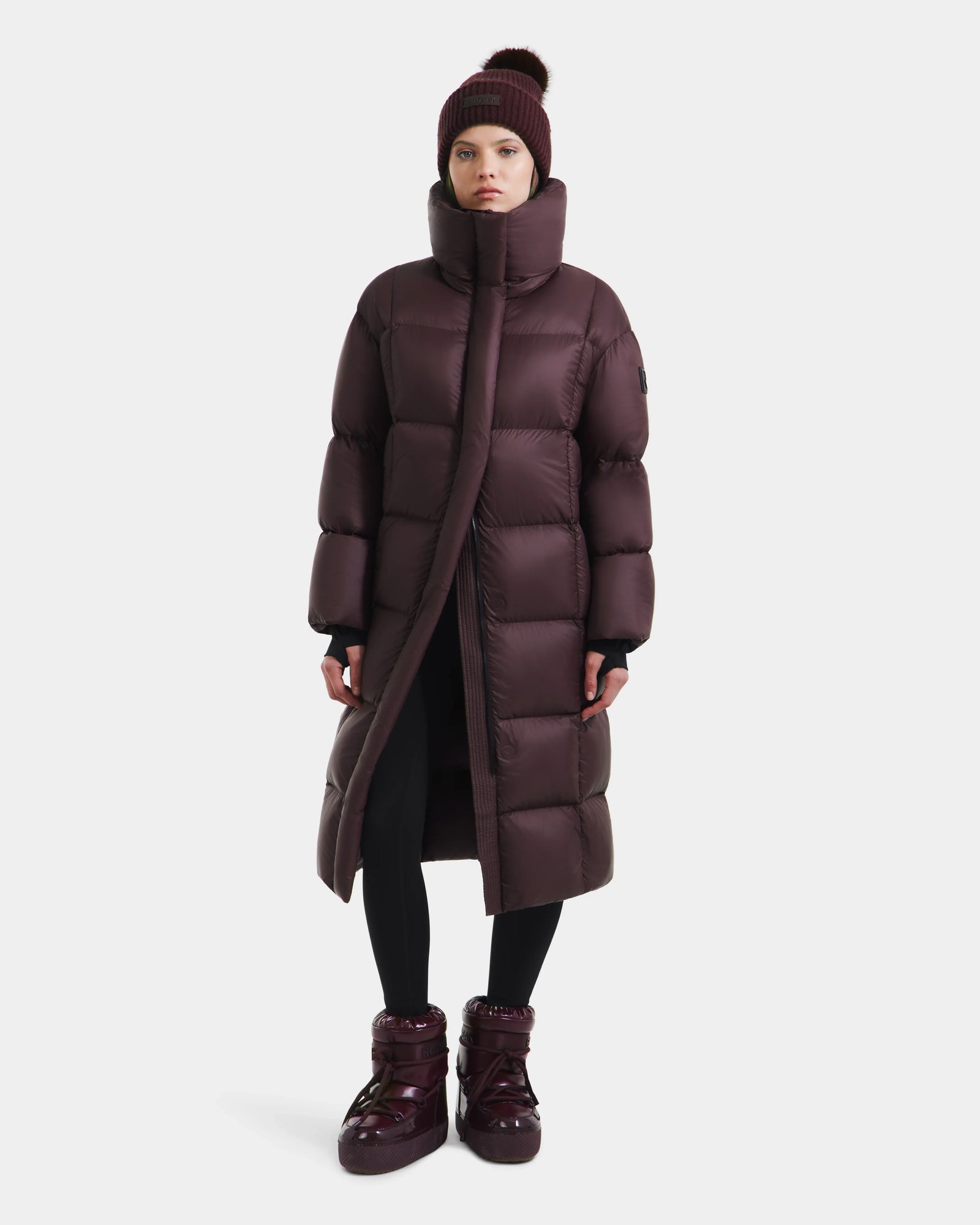 A person wearing the Rudsak SASHA - 8125686 Bordeaux long down puffer coat, matching boots, black leggings, and a maroon knit beanie stands against a plain white background.