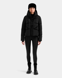 A person models the Rudsak STEFANIE M - L125526 Black puffer jacket, paired with black leggings, boots, and a fur hat, standing against a plain light gray background.