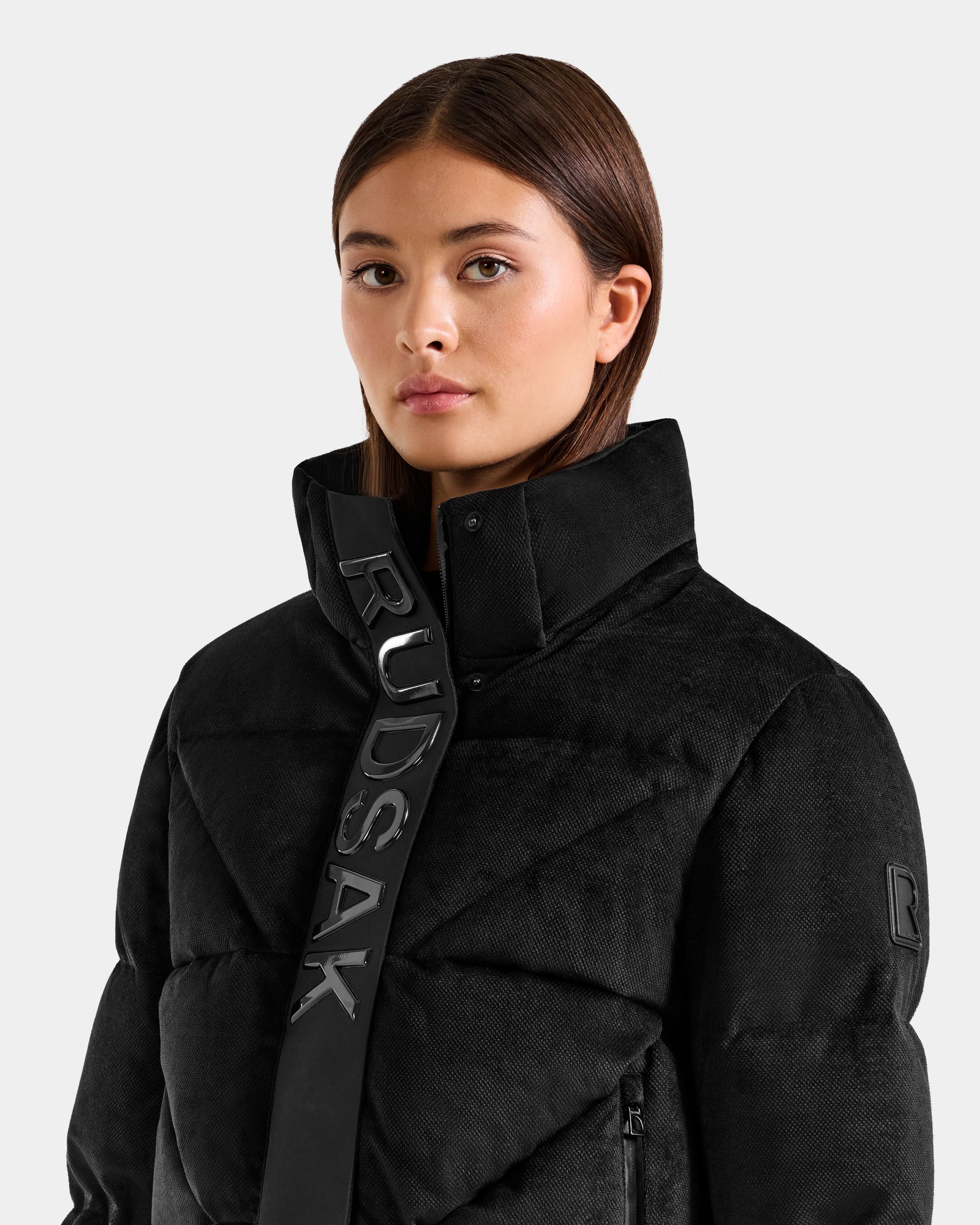 A woman with straight brown hair wears the Rudsak STEFANIE V - L125584 black down puffer jacket featuring a high collar with bold RUDSAK lettering, standing against a plain light gray background.
