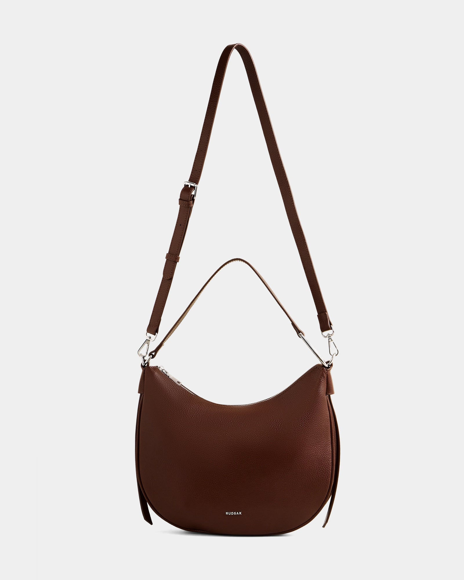 The Rudsak SYLVIA - L325515 Mocha is a women’s brown leather crescent shoulder bag with silver hardware, an adjustable crossbody strap, short handle, and small logo on the front, shown against a plain white background.