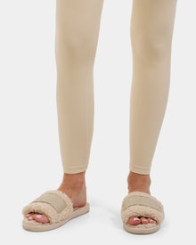 A person wearing beige leggings stands on a white background, showcasing Rudsak's TEDDY - L225517 Vanilla plush women's open-toe faux-shearling slippers. Only the lower legs and feet in cozy loungewear are visible.