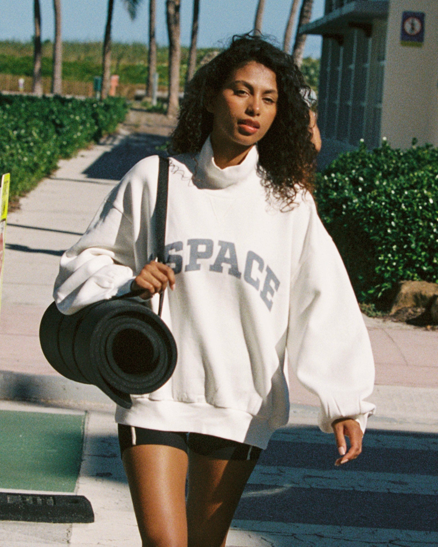 Outdoor lifestyle front view of the Crush Sweatshirt in cream, a cream quarter-zip pullover sweatshirt with "LSPACE" collegiate arch lettering, model carrying a yoga mat.